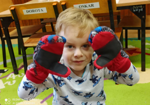 Chłopiec siedzi na dywanie. Pokazuje założone na dłoniach kolorowe rękawiczki ze Spiedermenem.